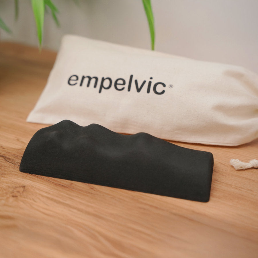 Schwarzes ergonomisches Sitzkissen für das Beckenbodentraining vor weißem Stoffbeutel mit Aufschrift 'empelvic' auf einem Holztisch