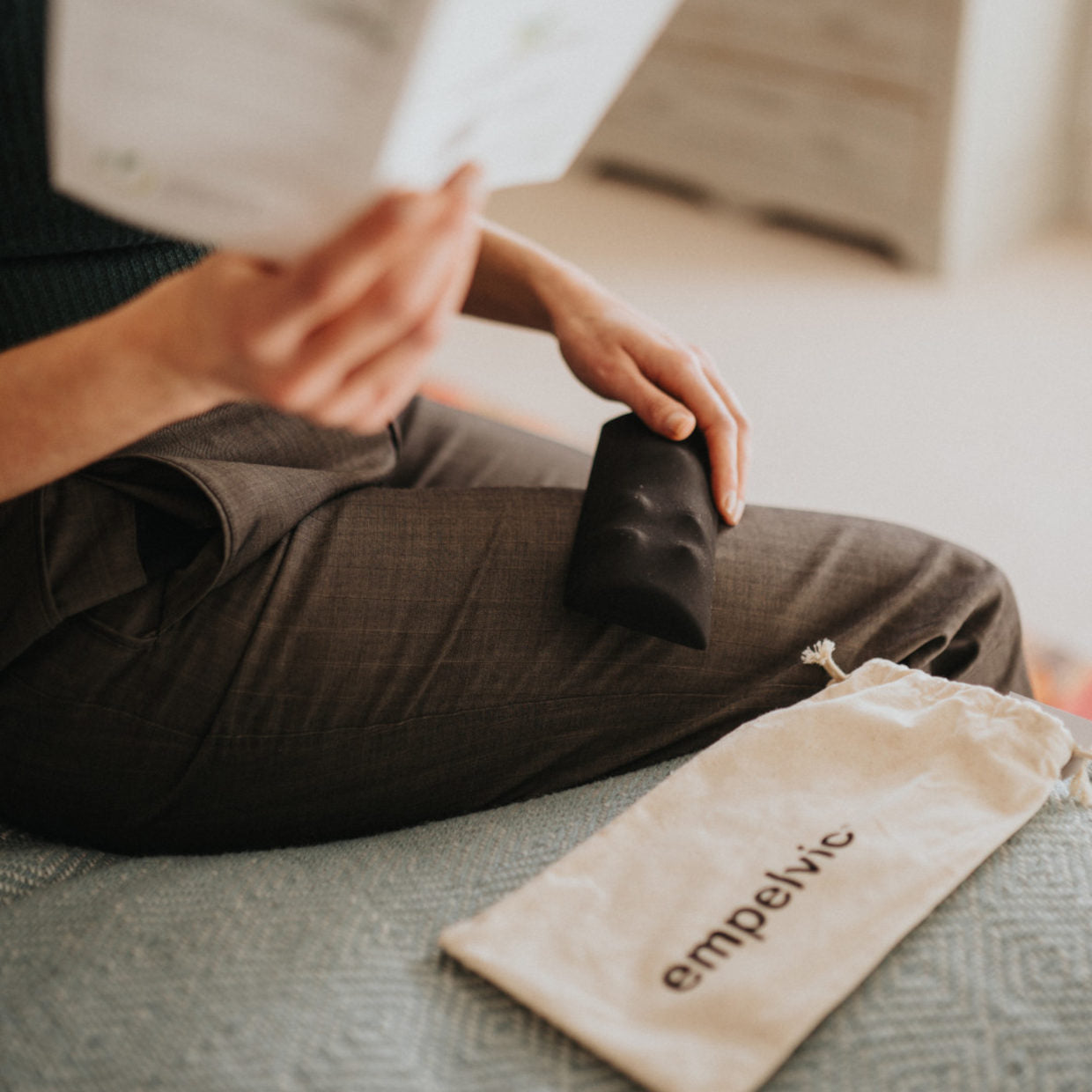 Person hält ein kleines schwarzes Empelvic Kissen auf dem Schoß und liest die Anleitung für das Beckenbodentraining, daneben liegt ein weißer Stoffbeutel mit dem Schriftzug &
